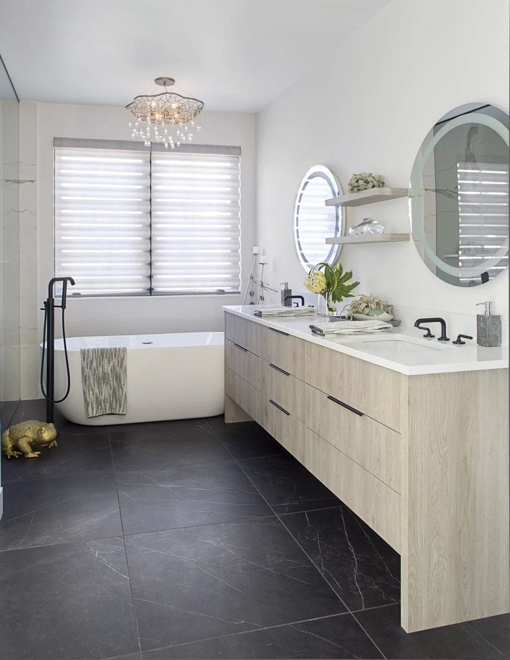Modern bathroom with a white bathtub near the window, black fixtures, dark tiled floor, light wood double vanity, round mirror, and decorative shelves, with a gold frog on the floor.
