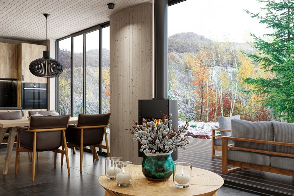 Modern living room with large windows showing a forested landscape with colorful autumn trees and a snow-covered ground outside. Inside, there are dining chairs, a round table with a floral centerpiece, a wood stove, and a cozy sitting area with a sofa and armchair.