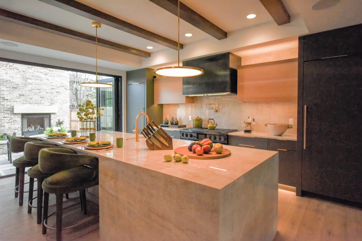 A modern kitchen with a large marble island, black and wooden cabinetry, and stainless steel appliances; soft lighting and decorative fruit on the counter.