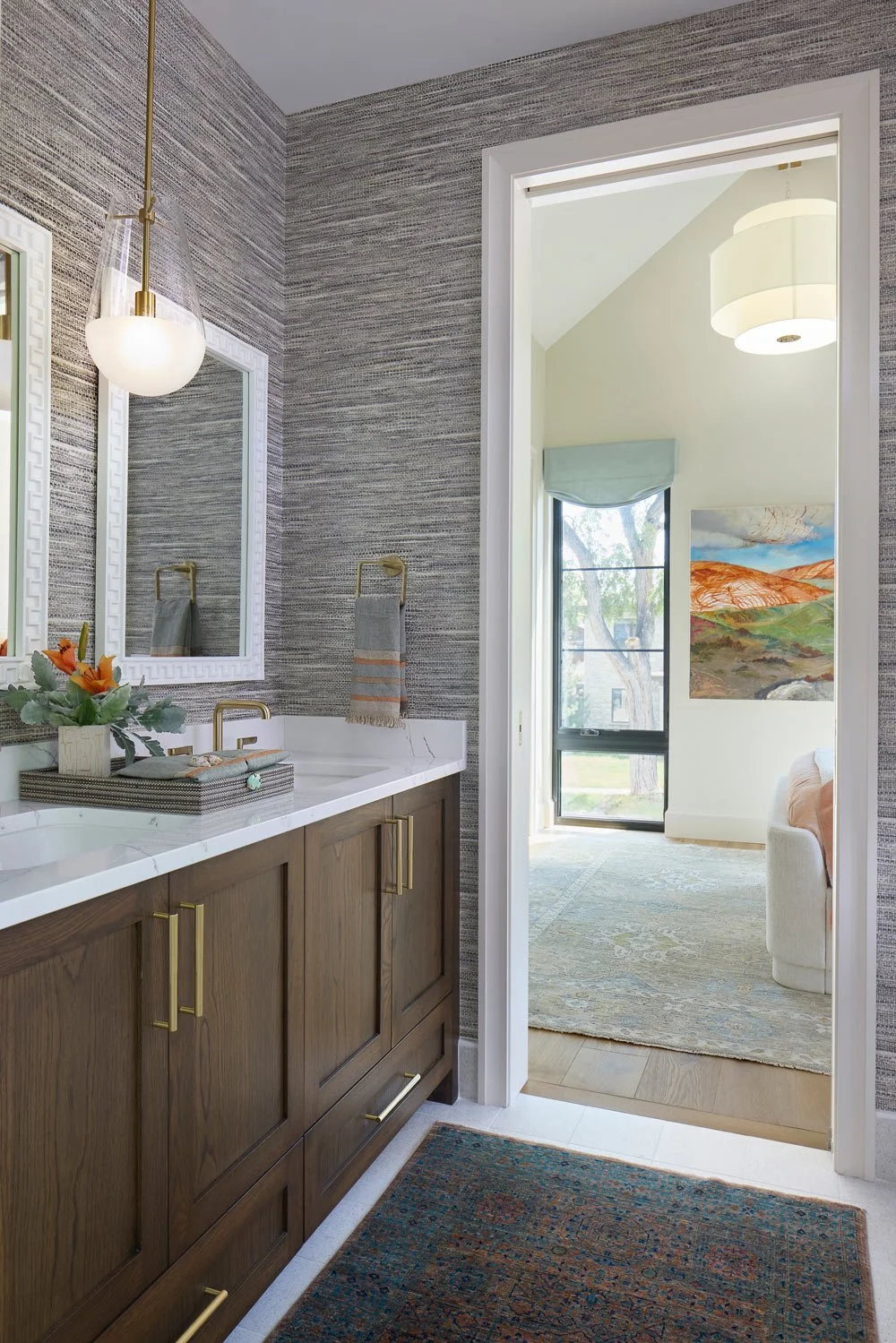 Bathroom with a dark wood vanity, marble countertop, large mirror, wall-mounted towels, decorative flowers, textured wallpaper, glass pendant light, and a doorway leading to a bright room with artwork, a window, and a sofa.