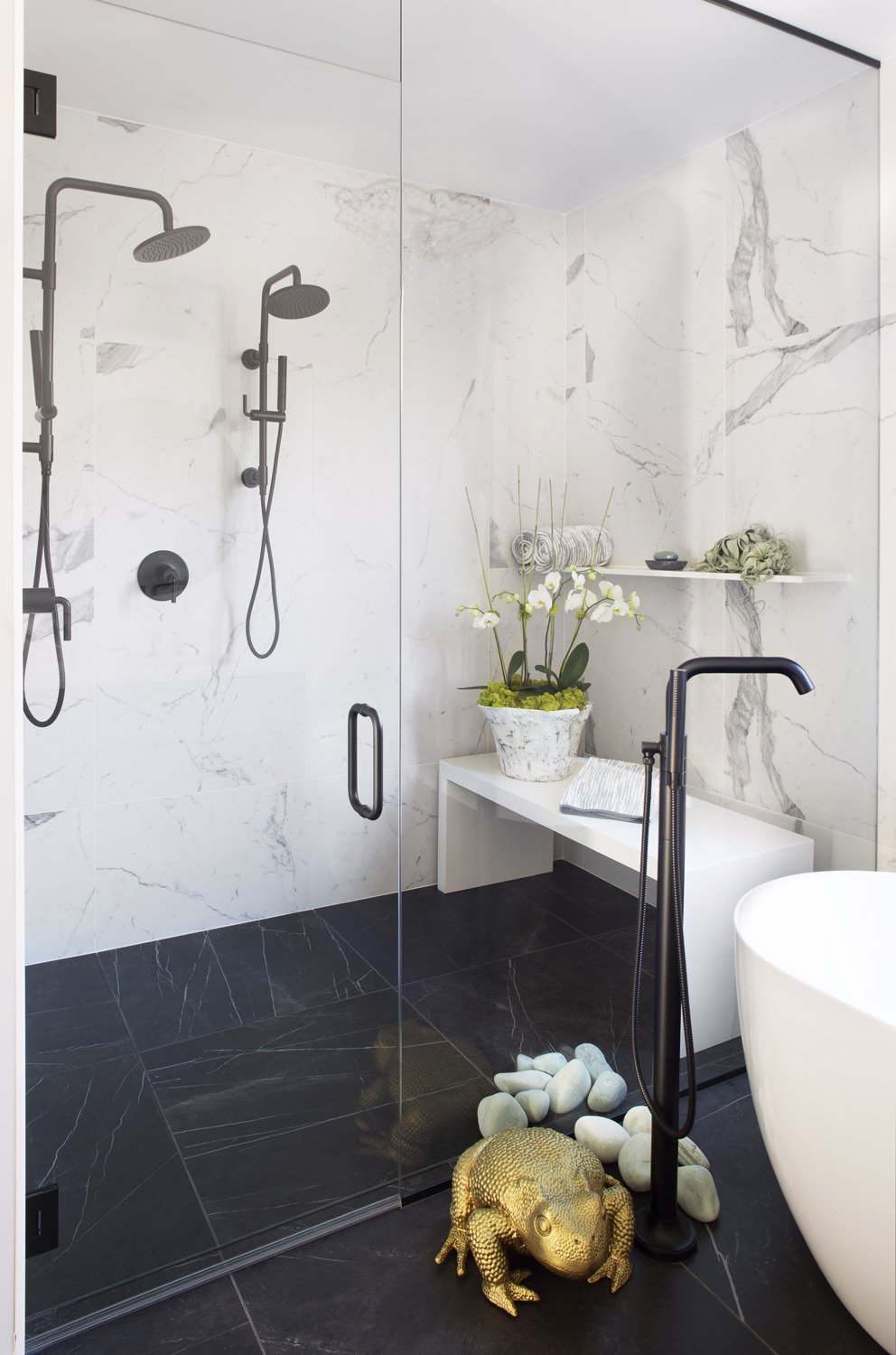 Modern bathroom with marble walls and black tiled floor, featuring a glass shower enclosure, black shower fixtures, a white bench with a potted orchid, a towel, and a decorative gold-colored toad sculpture surrounded by white stones, with a white freestanding tub partially visible.