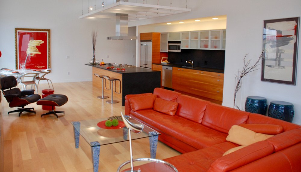 Open-concept living room and kitchen with orange leather sectional sofa, glass coffee table, modern art on the walls, and a kitchen island with bar stools.