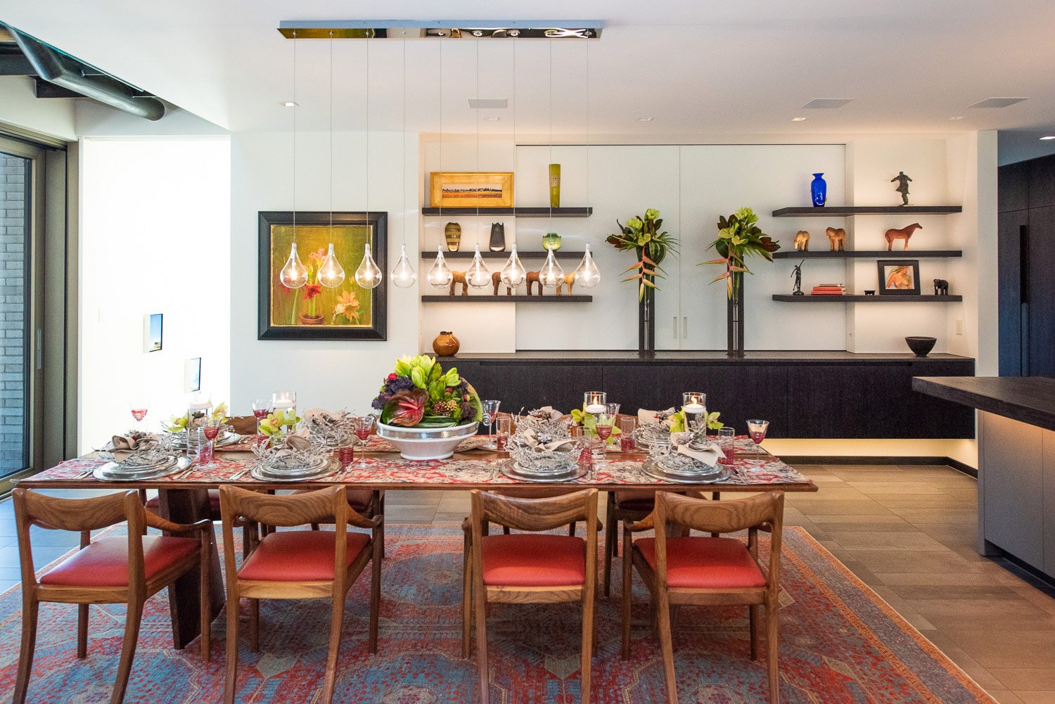 Dining room with a large table set for a meal, decorated with a floral centerpiece, surrounded by wooden chairs with coral cushions. The wall features white shelves with decorative items, and a large painting with bright colors. The room is well-lit with hanging glass teardrop-shaped light fixtures and natural light coming from sliding glass doors.