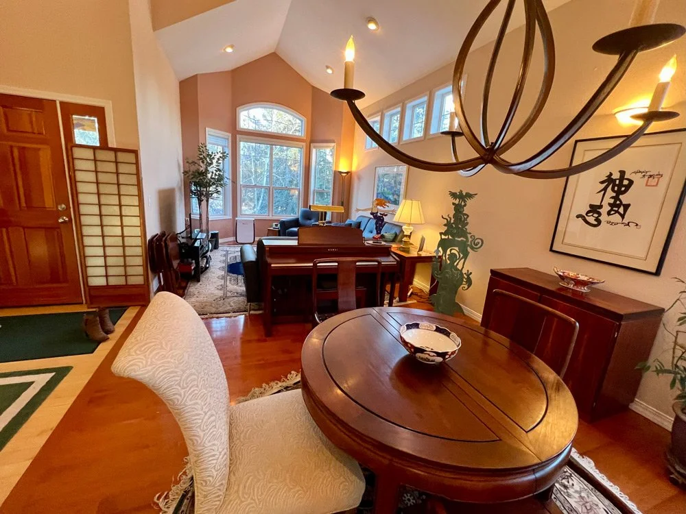 A bright living room and dining area with hardwood floors, featuring a round wooden dining table with a decorative bowl, a chandelier, a piano, seating area with a sofa, armchairs, lamps, large windows, and decorative art on the wall.