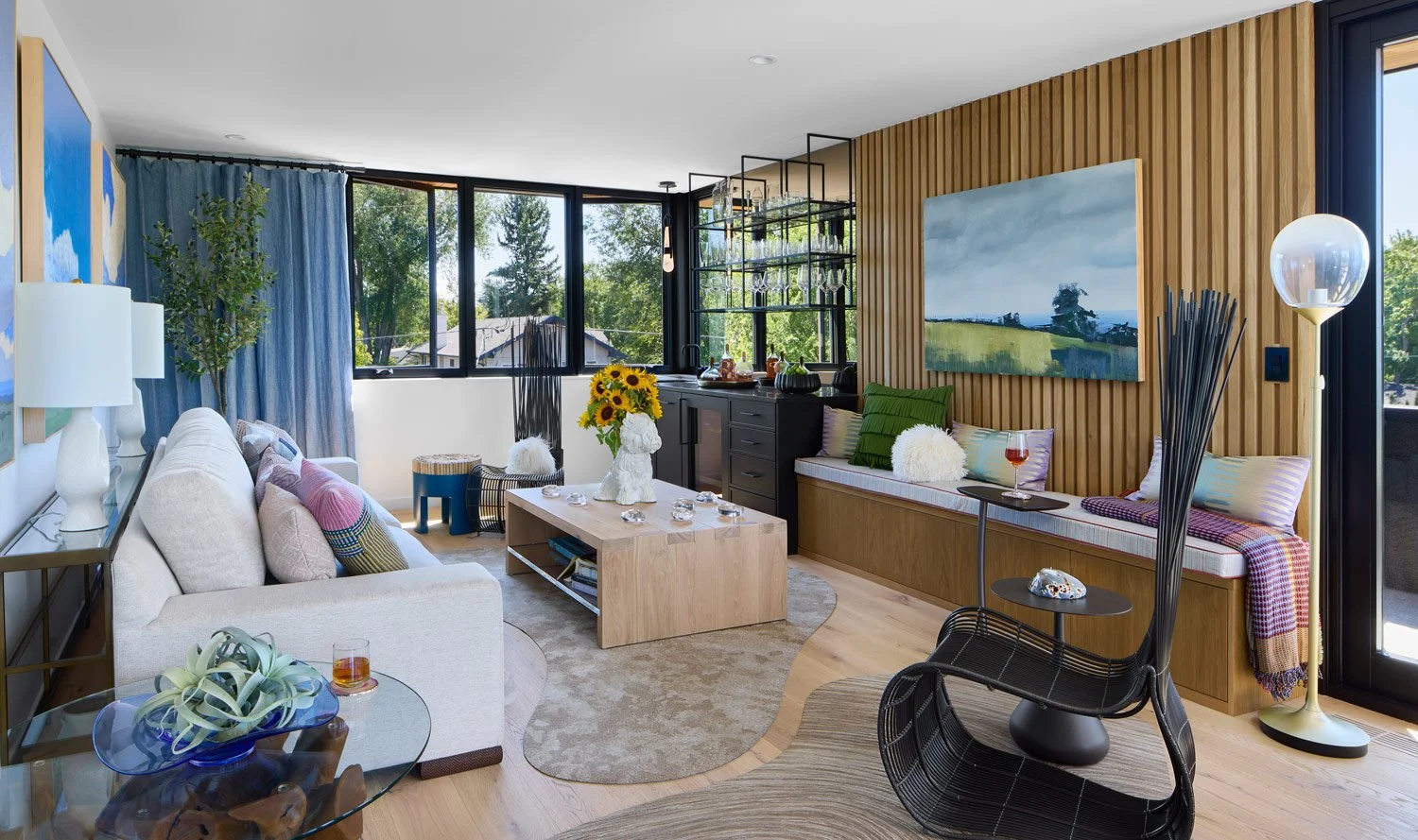 A modern living room with wooden paneling, a white sofa with colorful pillows, a round wooden coffee table, a built-in bench with pillows and a throw blanket, large window with blue curtains, and various decorative items including a painting, a lamp, and plants.