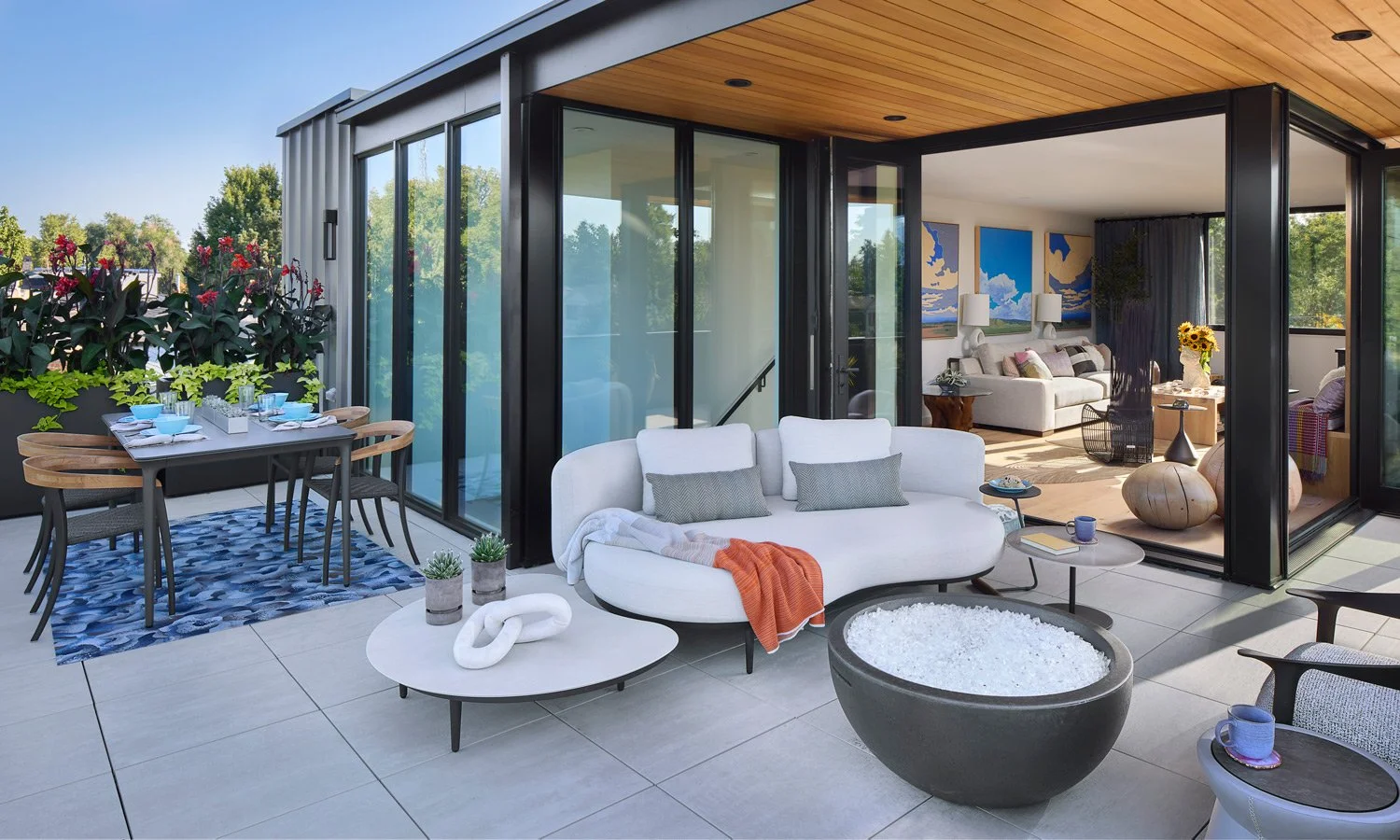 Modern outdoor patio with dining table, potted plants, and a white sofa. Inside, a living room with artwork, a large white sofa, and sunflowers. Sliding glass doors connect the patio and interior.