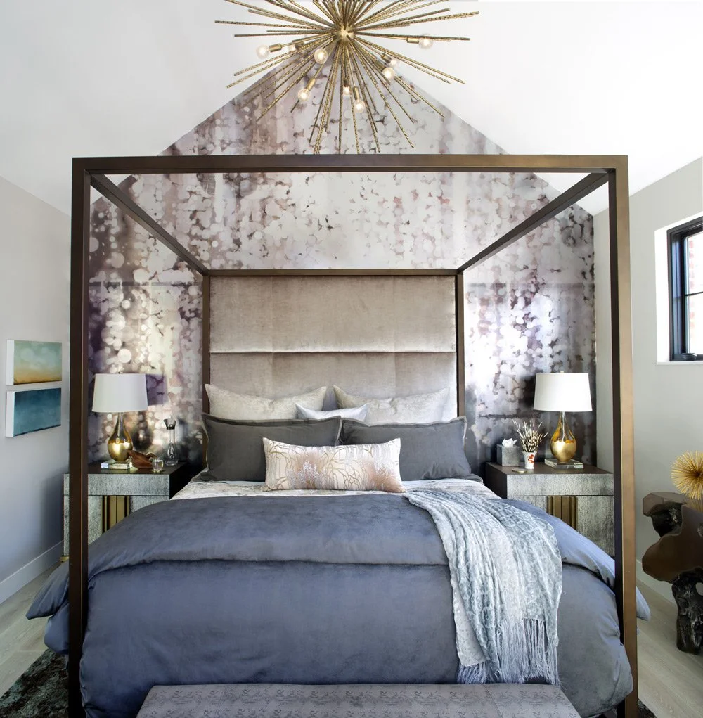 Modern bedroom with a four-poster bed, textured gray bedding, decorative pillows, and a metallic gold and glass bedside lamp on each side. The headboard is beige fabric, and a large metallic sputnik chandelier hangs from the ceiling. The walls have a marble-like wallpaper and a window on the right provides natural light.