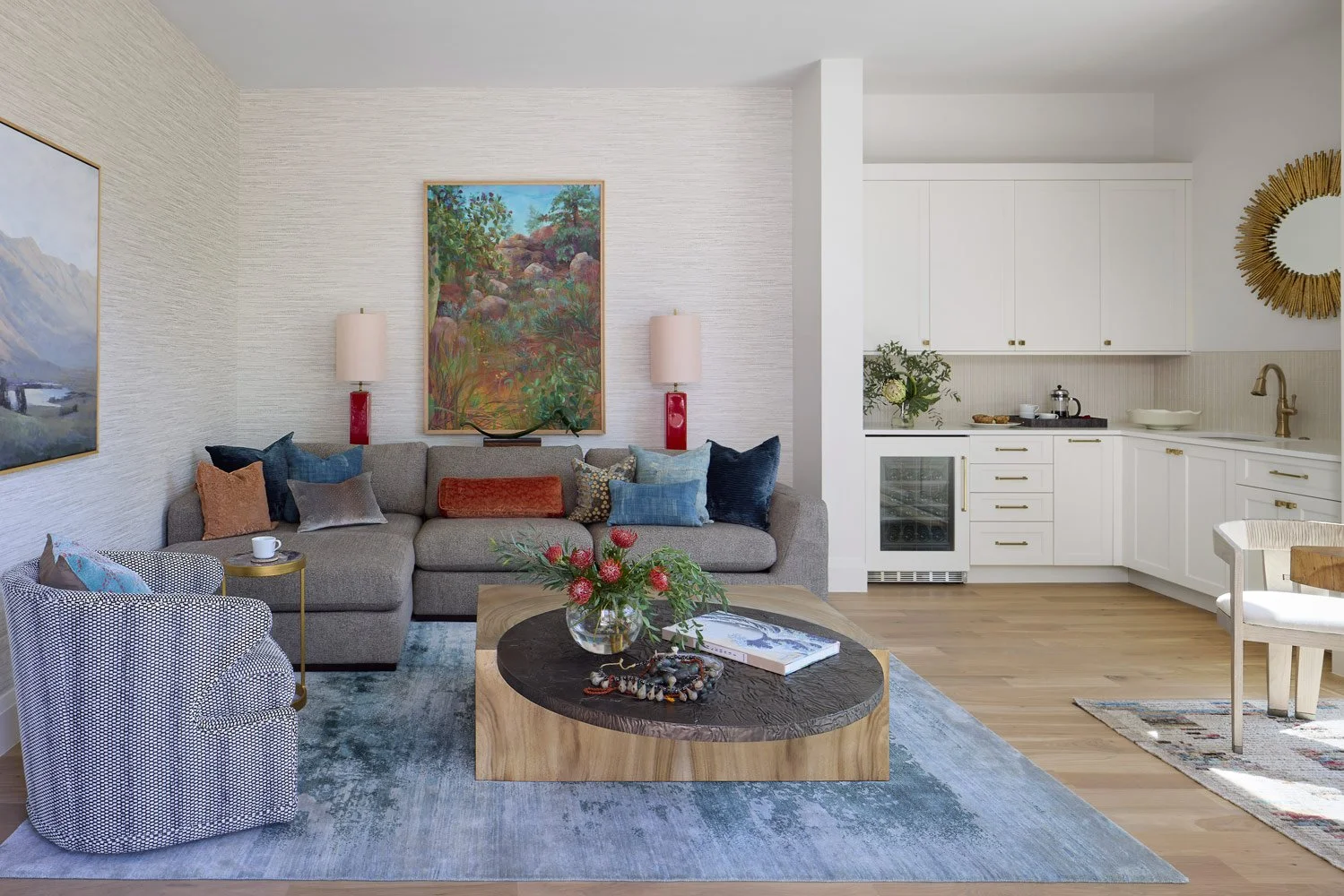 Living room with a gray sofa and patterned armchair on a blue rug, a wooden coffee table with a flower arrangement, two wall lamps, and framed landscape paintings in an open kitchen with white cabinets and gold hardware.