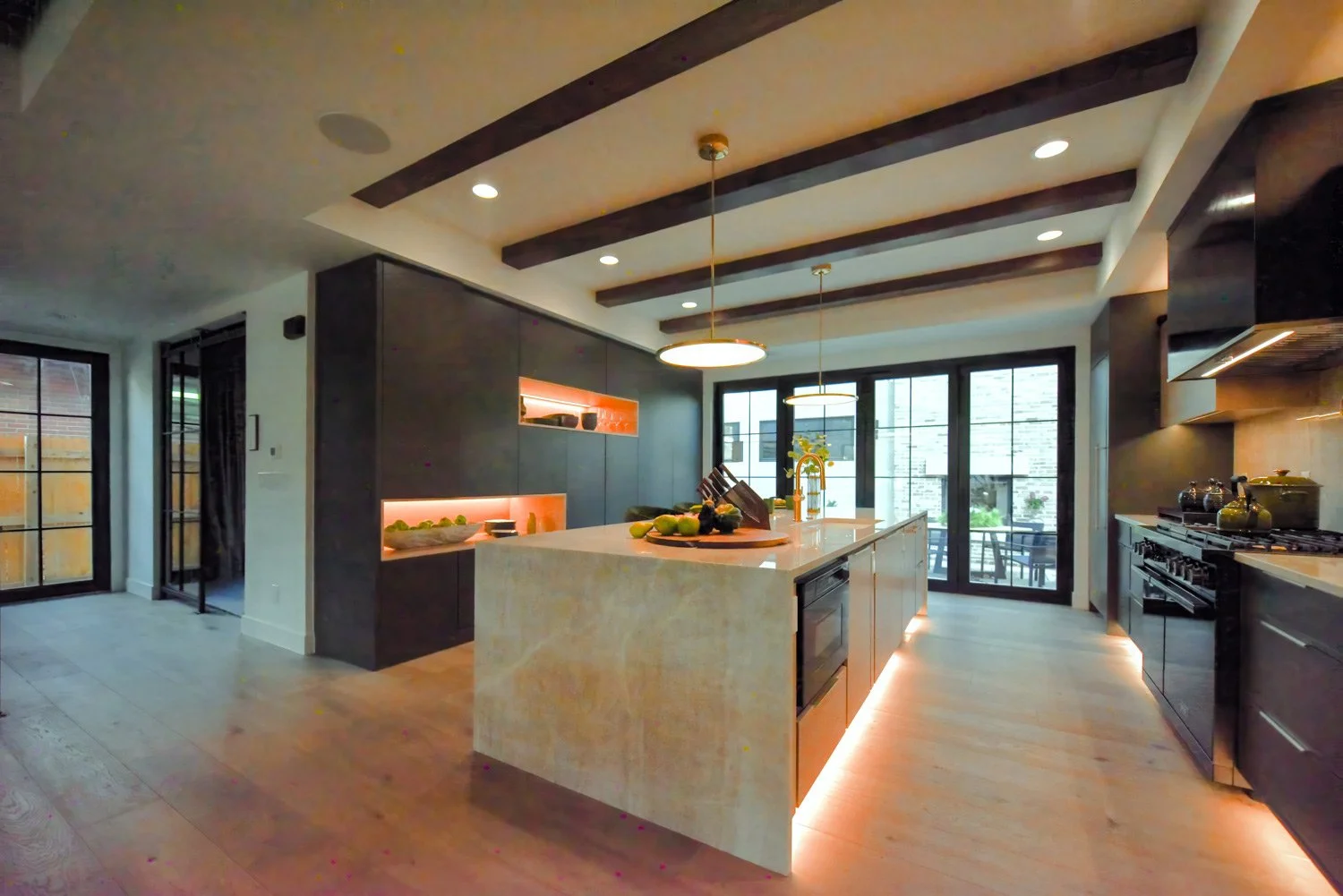 Modern kitchen with dark cabinets, a large central island with a light-colored stone countertop, pendant lights hanging from a ceiling with exposed dark wooden beams, and sliding glass doors leading to an outdoor patio.