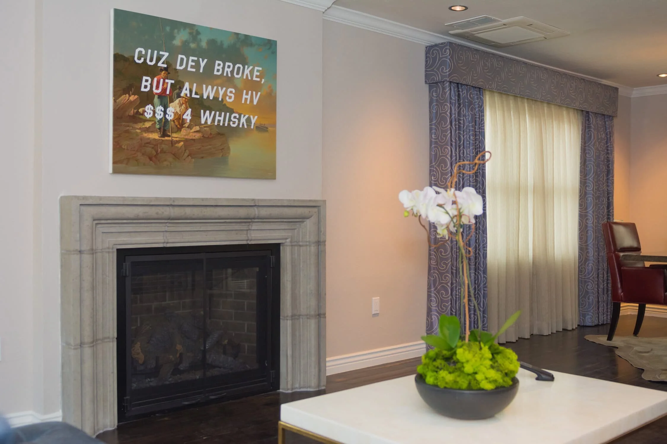 Living room with a fireplace, a large wall painting with humorous text about whiskey, and a window with blue patterned curtains. A white table with a flower arrangement in a gray pot is in the foreground.
