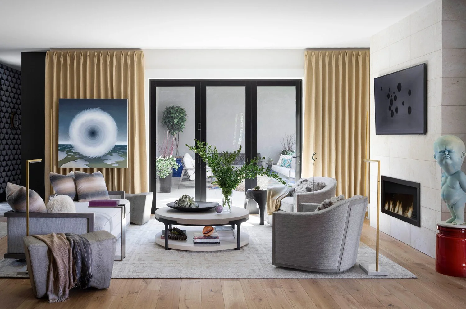 Modern living room with gray sofas, armchairs, a round coffee table, large gold curtains, a fireplace, and artwork on the walls, with sliding glass doors leading to a patio.
