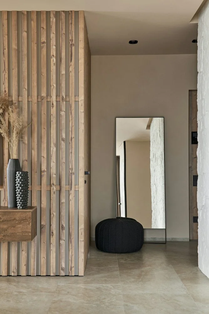 Indoor space with a wooden slat partition wall, a floating wooden shelf with vases, a large mirror, a stool, and a textured white wall.