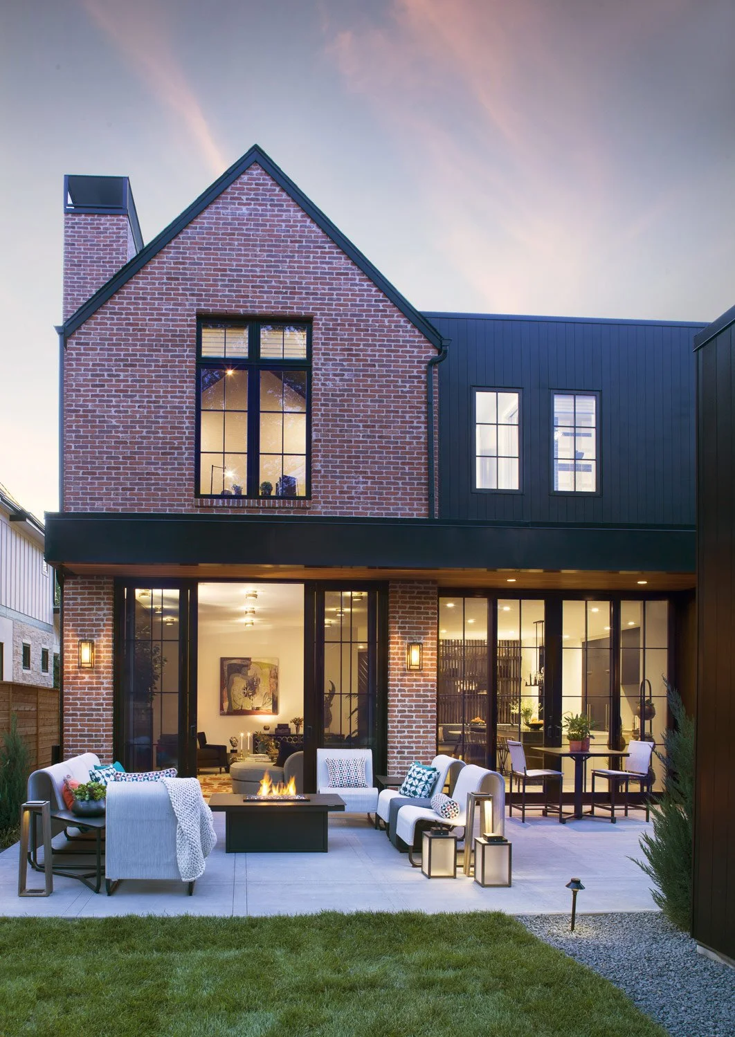 Modern two-story house with brick and dark siding exterior, large glass sliding doors opening to a patio with outdoor seating and firepit, overlooking a well-kept lawn.