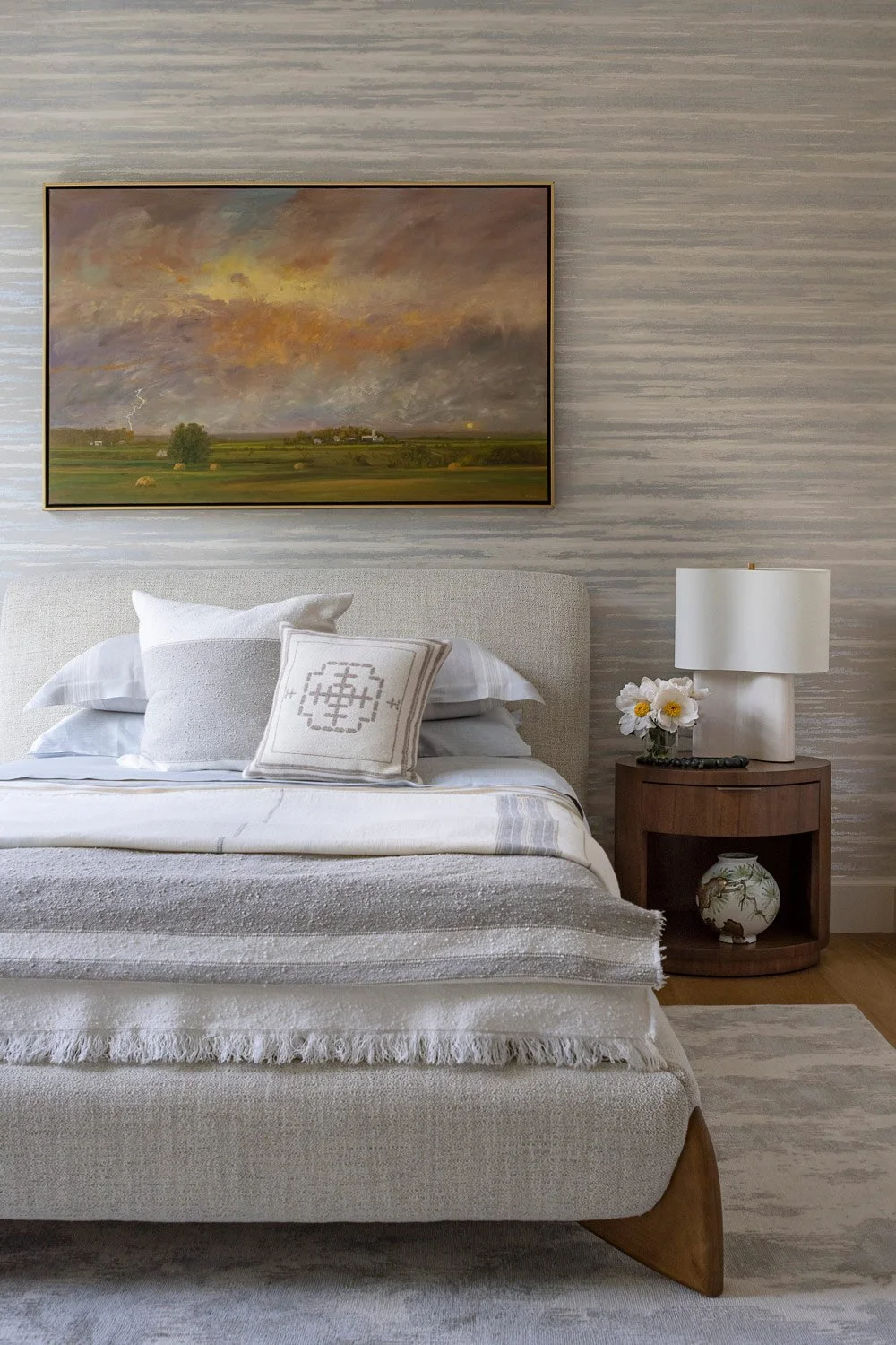 A bedroom with a neutral-toned upholstered bed, pillows, a side table with a vase of flowers, a white lamp, and a landscape painting of a stormy sky over a green field hanging above the bed.