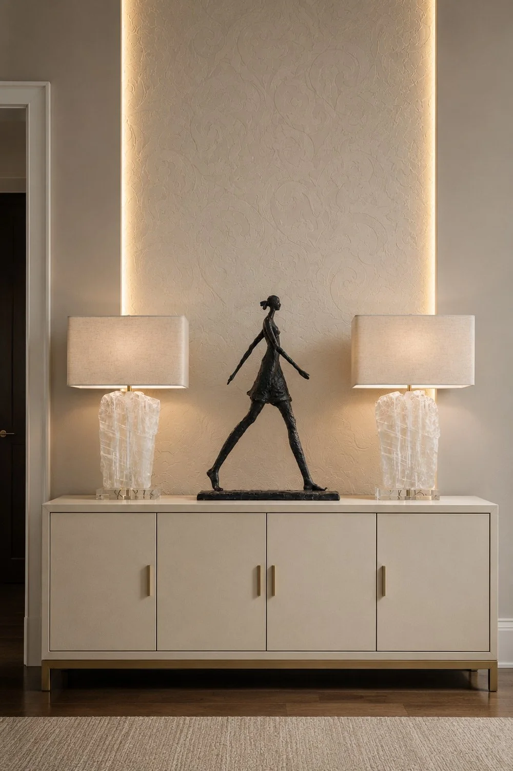 Decorative interior with a white sideboard, two table lamps with white shades and textured bases, a sculpture of a walking woman in black, and warm lighting accent on textured wall background.