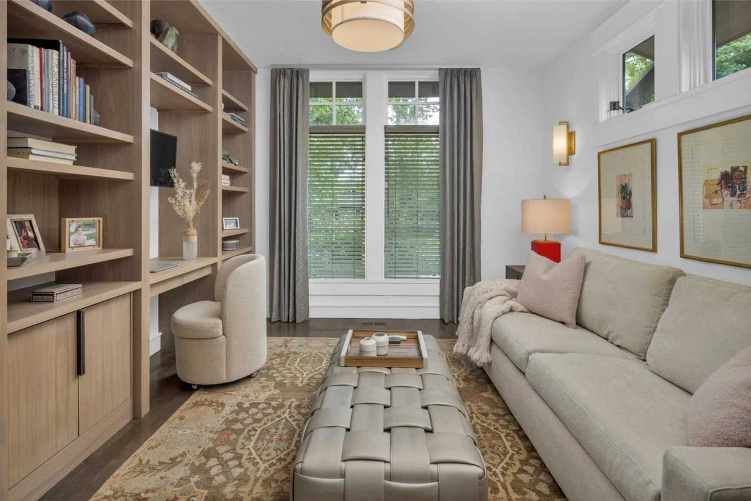 Living room with beige sofa, armchair, wooden bookshelf, window with blinds and curtains, patterned area rug, lamps, framed art on wall, and decorative tray on tufted ottoman.