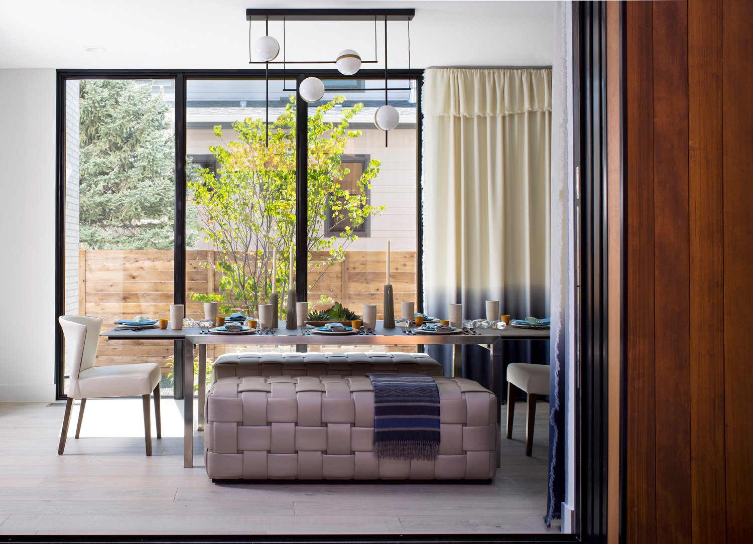 A modern dining room with a large window, a white table set with dishes, glasses, and tall vases with candles, two white chairs, a woven pink ottoman with a blue blanket, and a view of a wooden fence and trees outside.