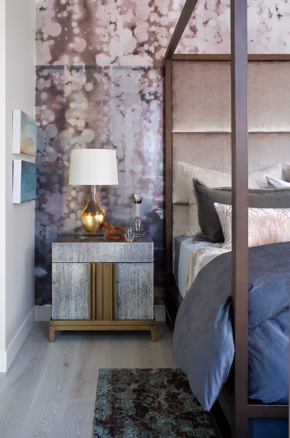 A bedroom corner with a side table, a lamp with a white shade, a glass carafe, and a small carved figure, next to a bed with a beige headboard and decorative pillows. The wall behind has a pink and white bokeh pattern.