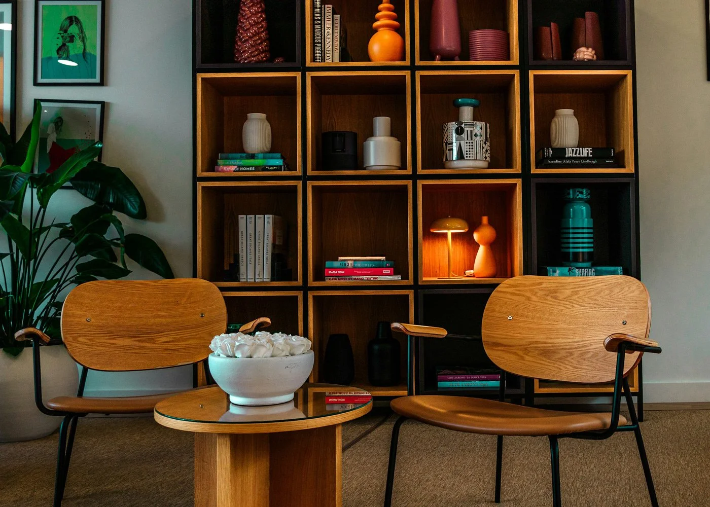 Living room corner with a wooden shelving unit filled with decorative items and books, two wooden chairs with black metal frames, a small round table with a white bowl containing tissue, a large green plant, and framed artwork on the wall.