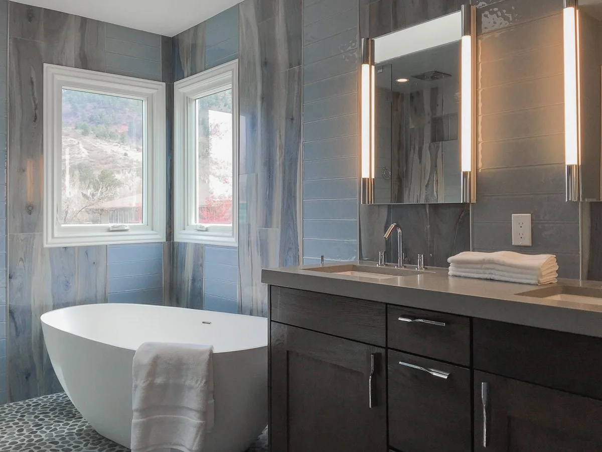 Modern bathroom with a freestanding bathtub, a vanity with a mirror and lighting, and two windows showing an outdoor view.