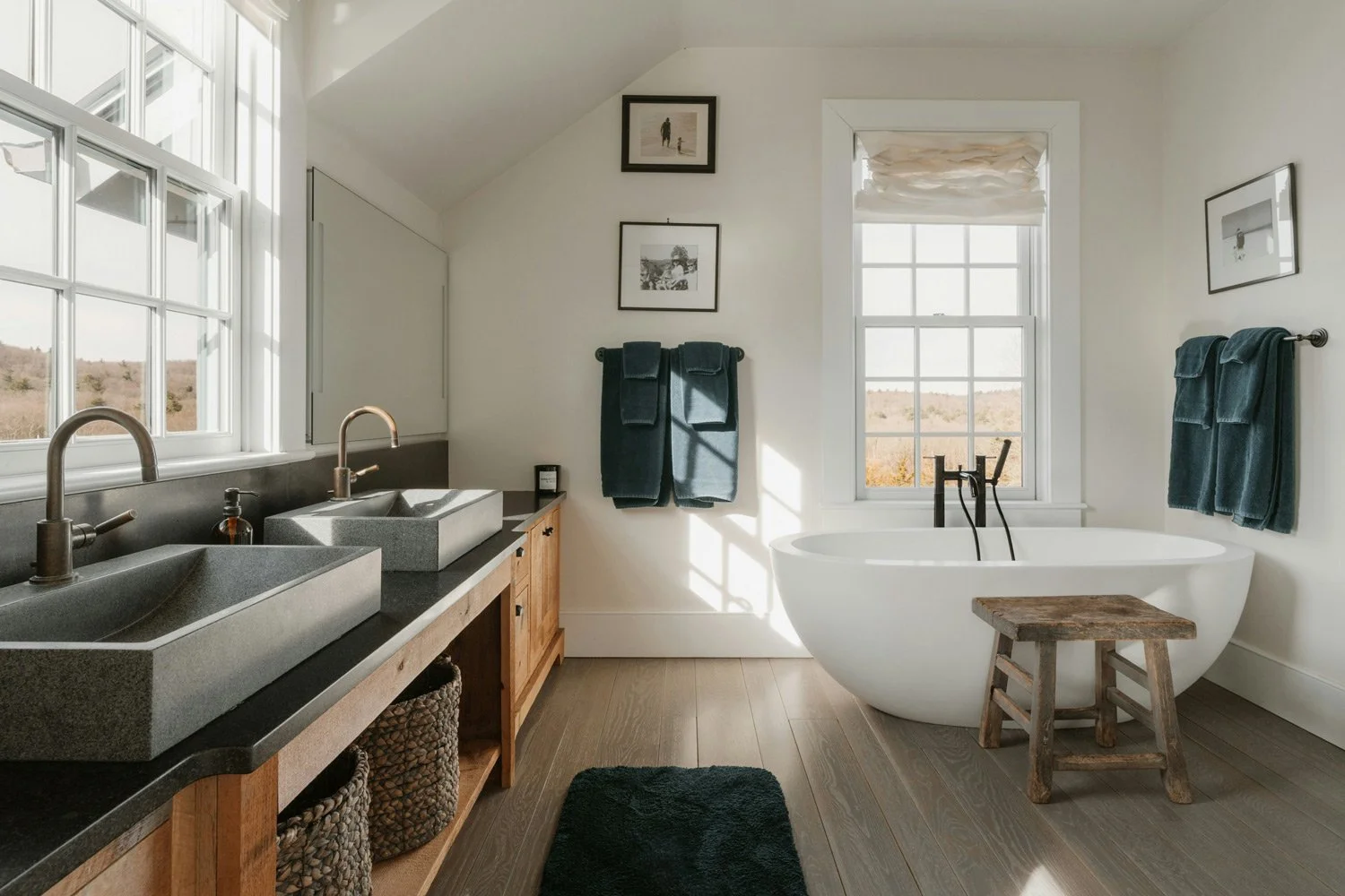 Bright bathroom with double sinks, black countertop, wooden cabinet, two wall-mounted towel bars with dark towels, freestanding bathtub near a large window, small wooden stool, framed artwork, and a black rug.