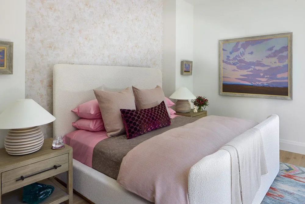 A bedroom with a white upholstered bed, pink and beige pillows, a pink and brown blanket, and bedside tables with lamps and decorative items. There is a framed landscape painting on the wall.