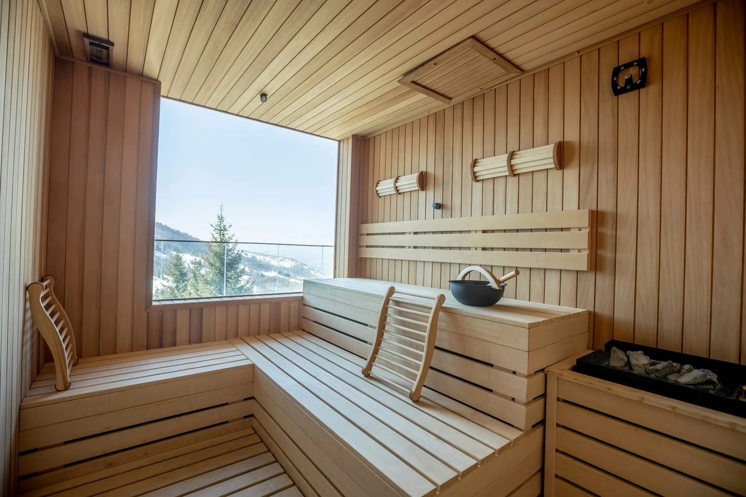 Interior of a traditional Finnish sauna with wooden benches, a sauna water bucket, and a heater with rocks, overlooking a snowy mountain landscape through an open window.