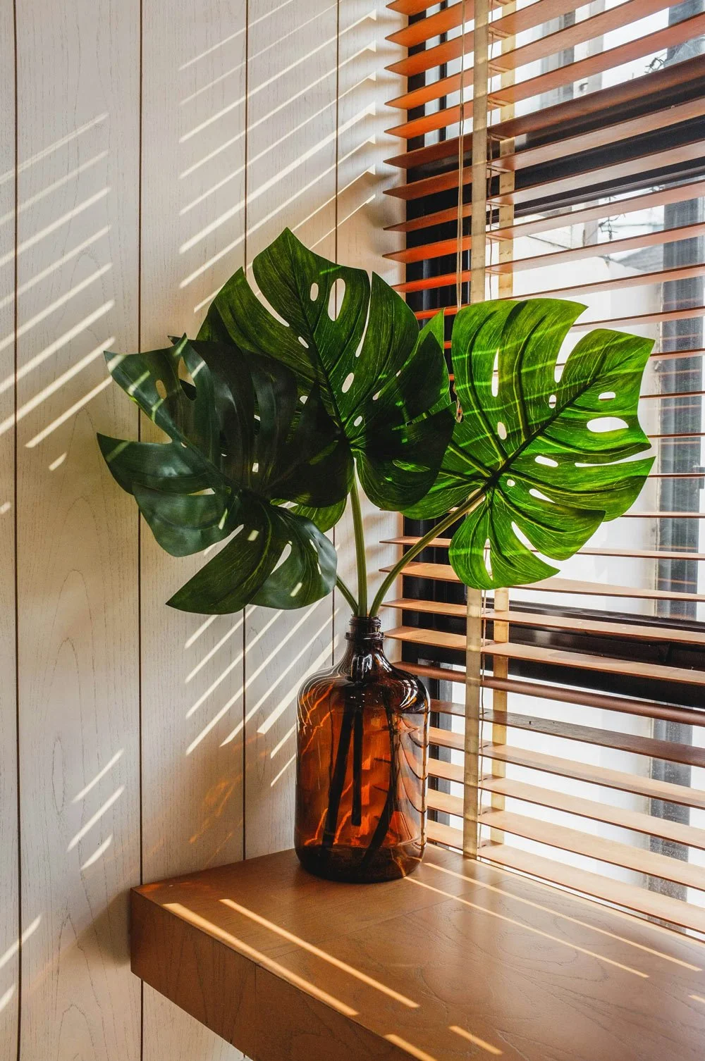 A green tropical monstera plant in an amber glass vase placed on a wooden shelf next to a window with wooden blinds, casting shadow lines on a light-colored wall.