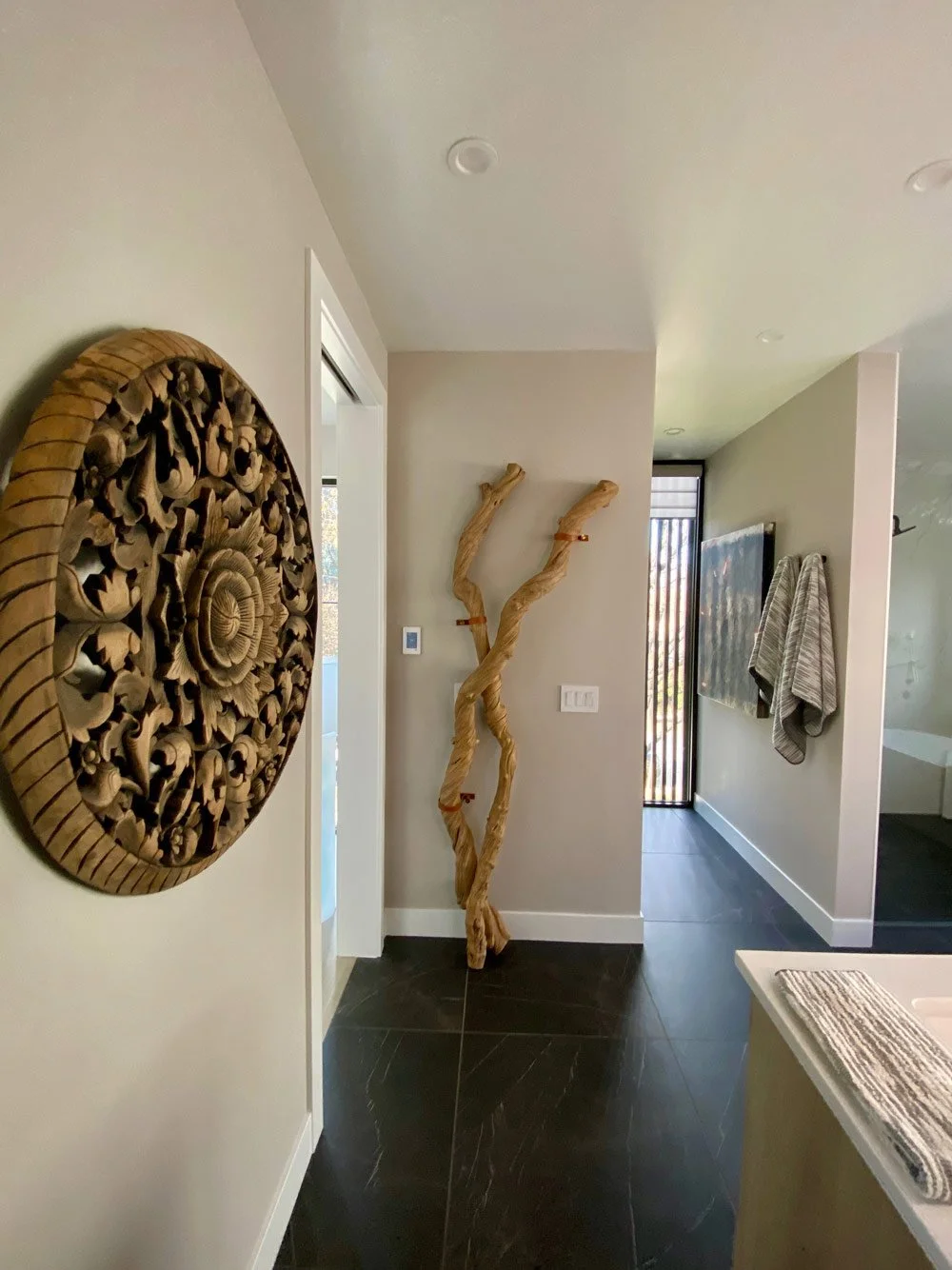Interior of a modern home with a carved wooden wall art on the left, a twisted wooden sculpture in the center, and a wall-mounted television with towels on a rack on the right.