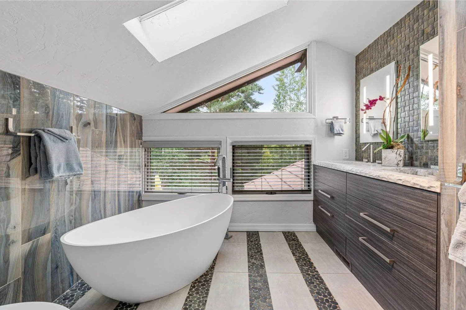 Modern bathroom with a white freestanding bathtub, large windows with blinds, a dark wood vanity with a marble countertop, a mirror, and a potted plant with pink flowers.