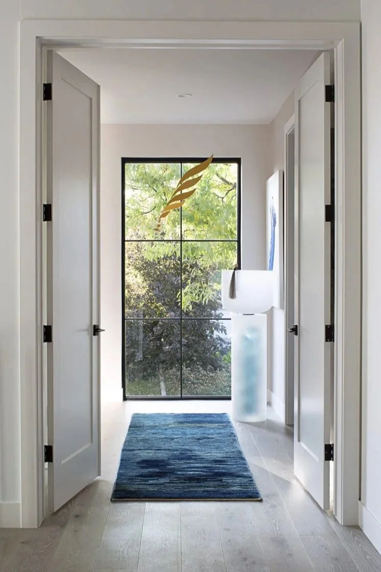 View through an open doorway into a bright room with a large window showing greenery outside. A blue rug is on the floor, and there is a tall white sculpture or stand near the window.
