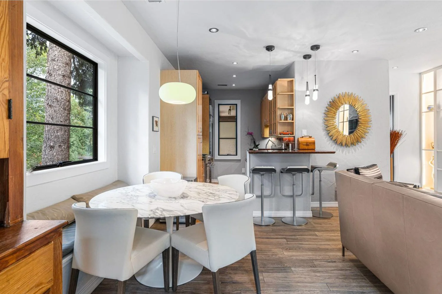Open-concept living area with dining table, bar counter, and kitchen. Large window showing trees, white walls, wooden accents, and modern lighting fixtures.
