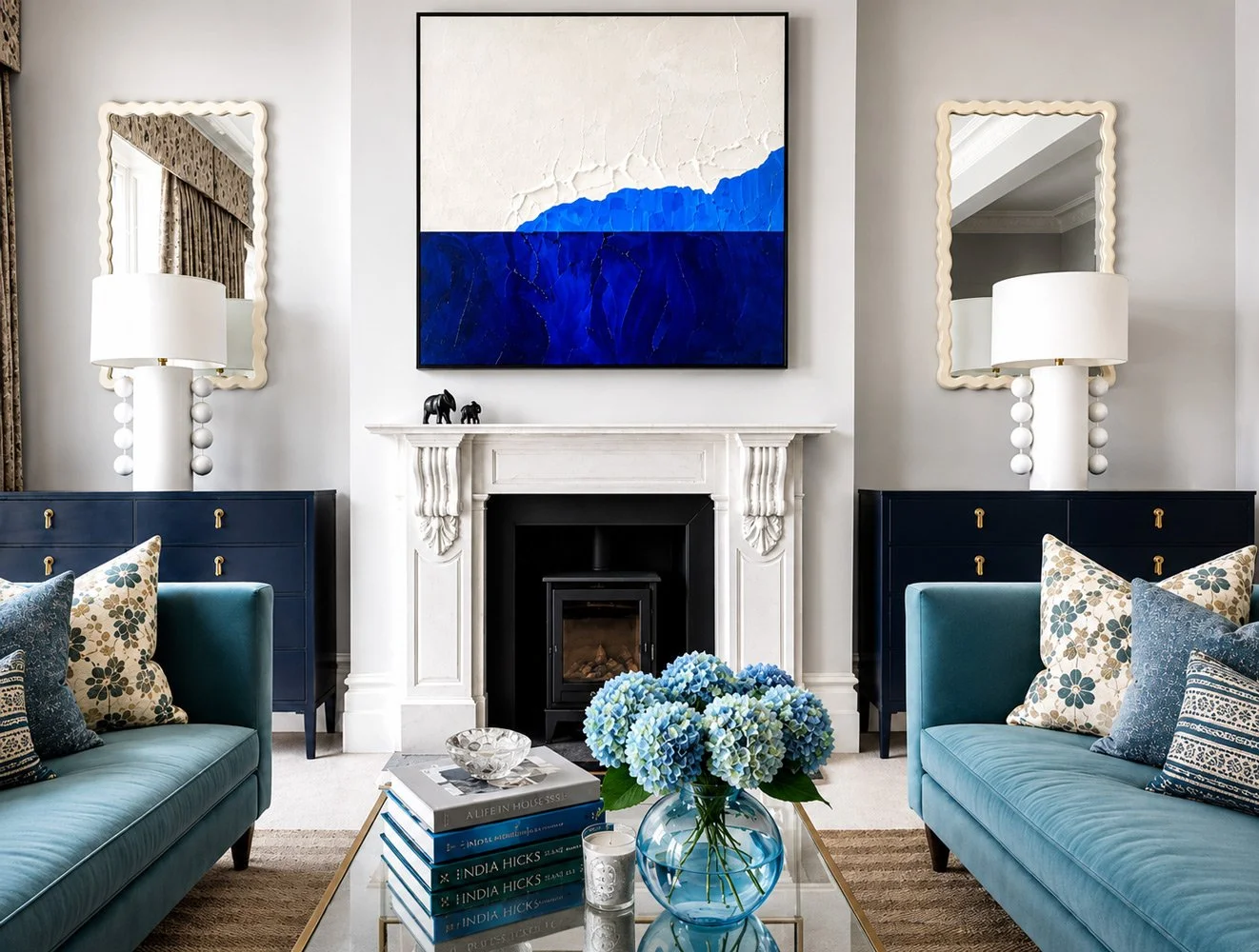 Living room with two blue sofas, a white fireplace, two matching mirrors, table with books and a blue hydrangea flower in a glass vase, and artwork with blue and white textured sections above the fireplace.
