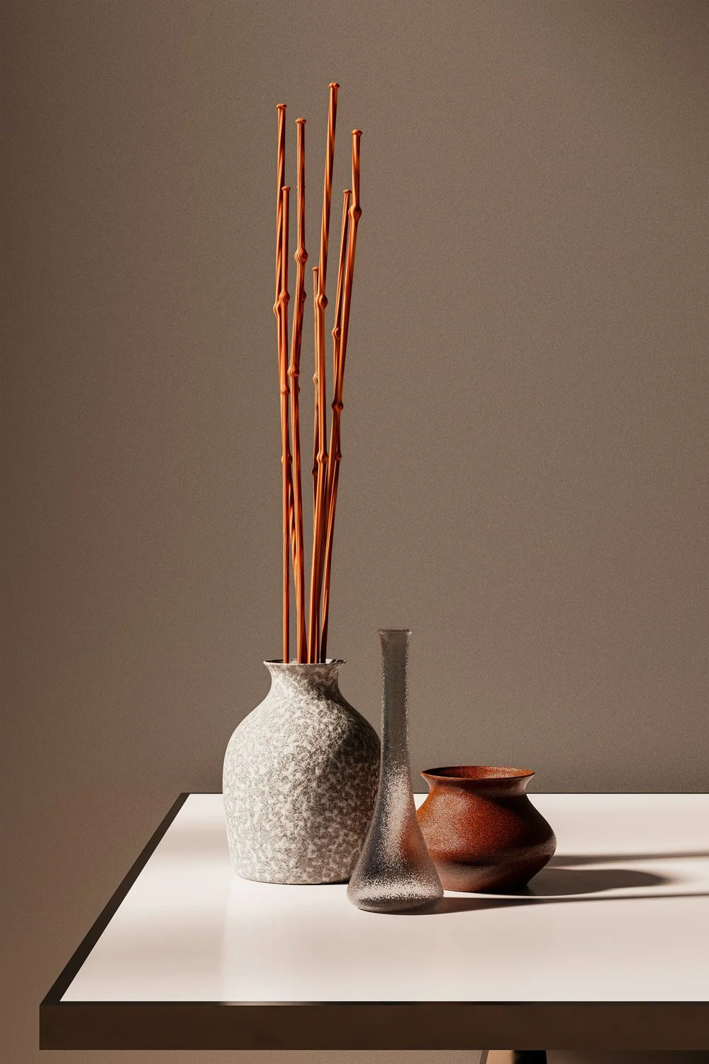 Three decorative vases, with the tallest holding long, brown, bamboo sticks, placed on a white table against a plain wall.