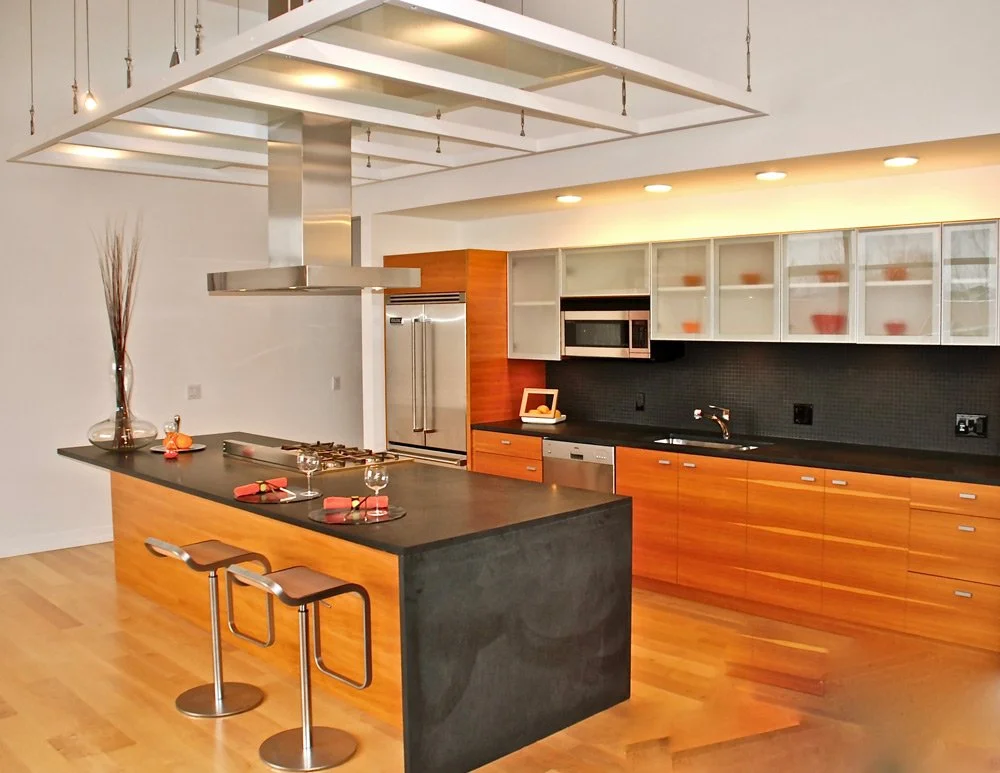Modern kitchen with black countertops, wooden cabinets, glass-front upper cabinets, built-in microwave, stainless steel refrigerator, and a kitchen island with two stools and place settings.