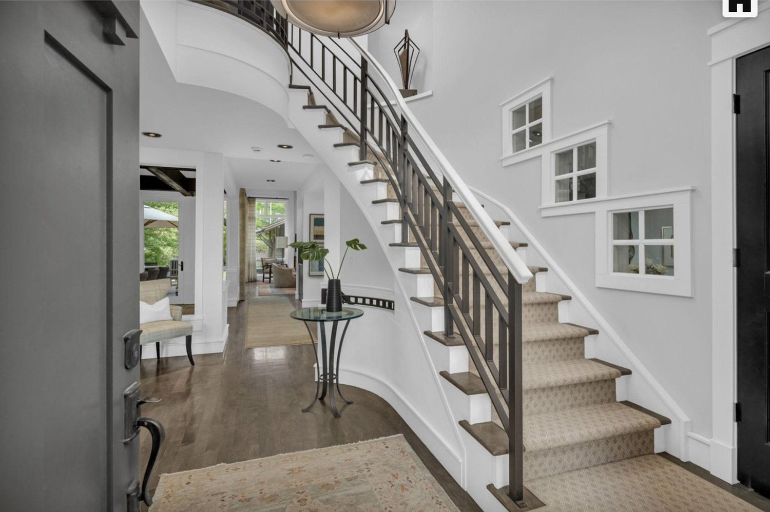 Entryway of a house with a curved staircase, dark wood railing, beige carpeted steps, white walls with trim, small round table with a black vase and plant, hardwood floor, and open interior with living and dining areas visible in the background.