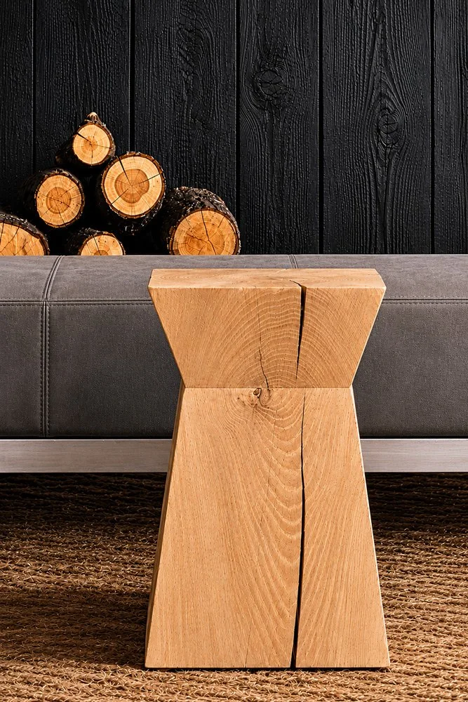 A wooden side table with a modern, geometric design placed in front of a gray sofa, with a black wooden wall and stacked logs in the background.