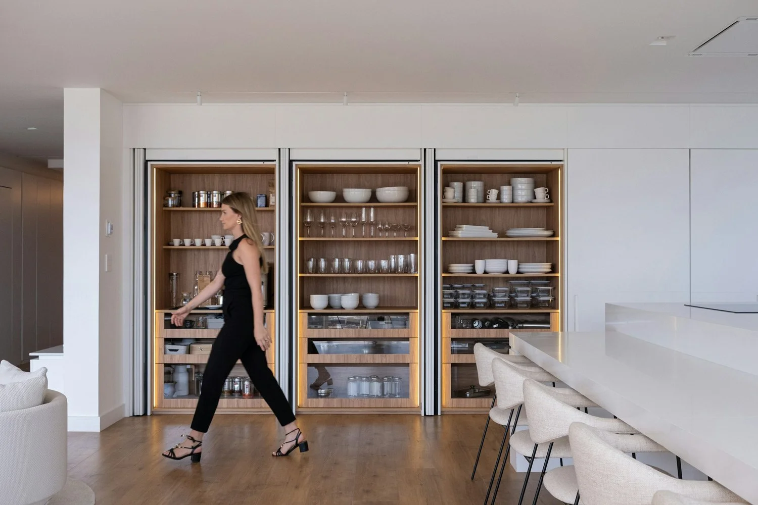 A woman in a black sleeveless outfit and black high-heeled sandals walks past a modern kitchen with wooden shelves filled with white dishes and glasses, and white chairs along a white island.