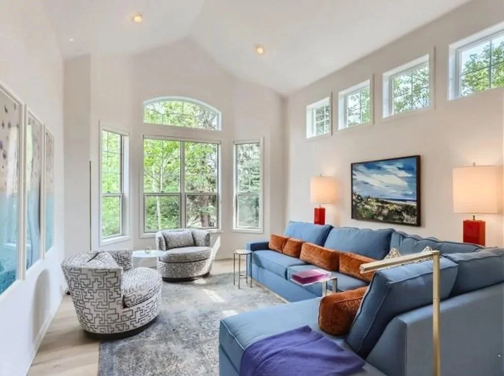 Bright living room with white walls, large windows with greenery outside, a blue sectional sofa with orange and patterned throw pillows, two patterned accent chairs, two table lamps with red bases, a wall-mounted TV, and a gray area rug.