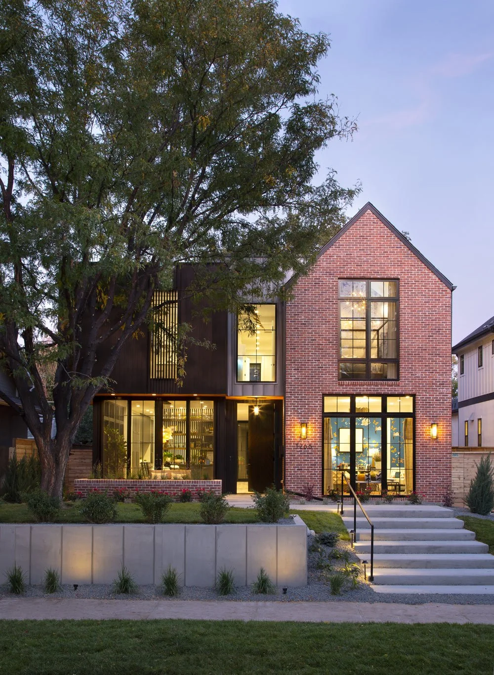 A modern two-story house with brick and black exterior, large front windows, steps leading up to the entrance, and a large tree in the front yard during evening.