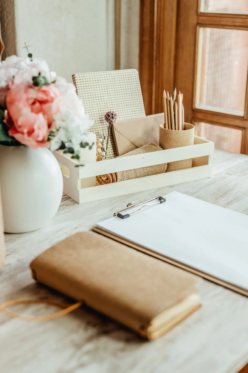 A tidy wooden desk with a vase of pink and white flowers, a white organizer holding beige foliage, a rolled towel, a paper in an envelope, a clipboard with blank paper, and a closed brown leather notebook.