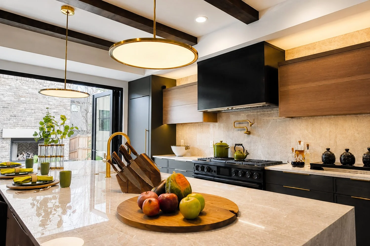 Modern kitchen with black and wooden cabinets, beige stone backsplash, and a marble countertop island. The island has apples and a cutting board. There are green bowls, a kitchen faucet, and a stove with a green kettle. Two round pendant lights hang from the ceiling, and a sliding glass door leads outside to a backyard with a brick wall.