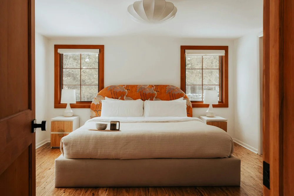 A bedroom with a bed, two nightstands with lamps, two windows with blinds, and a ceiling light fixture.