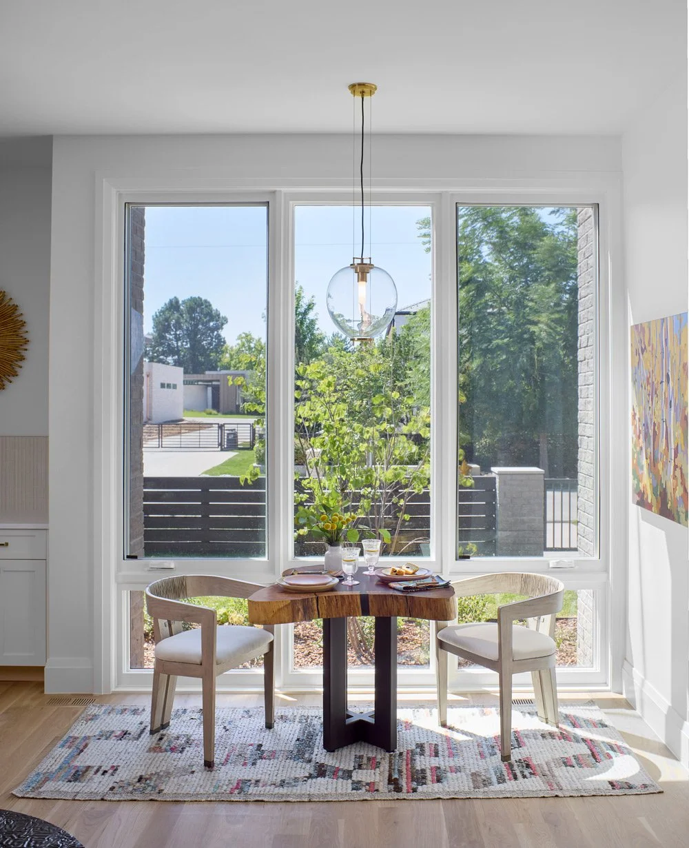 A bright dining nook with two white chairs, a small wooden table set for two, and large windows showing a yard with greenery outside.