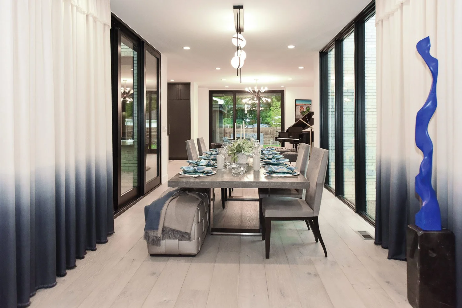 Modern dining room with a large wooden dining table set with plates, glasses, and a flower centerpiece, surrounded by beige chairs, with a black grand piano in the background, glass sliding doors, and contemporary lighting fixtures.