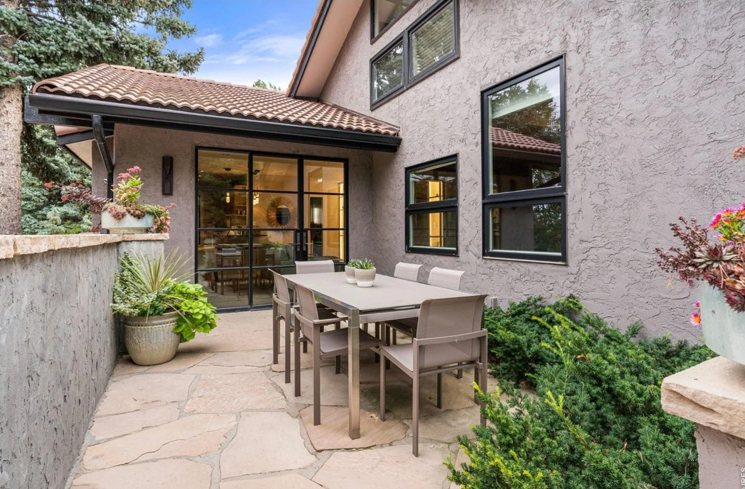 Outdoor patio with a dining table and chairs, surrounded by potted plants and greenery, attached to a house with a stucco exterior and black-framed windows.