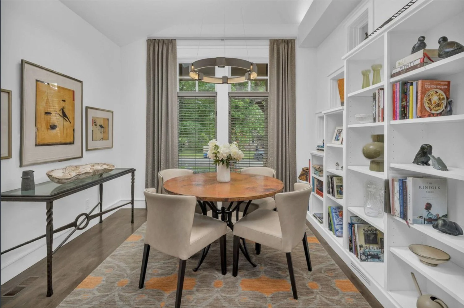 A dining room with a round wooden table, six beige upholstered chairs, a white bookshelf filled with books and decorative objects, a glass console table with art pieces, artwork on the walls, a large window with beige curtains, and a modern chandelie