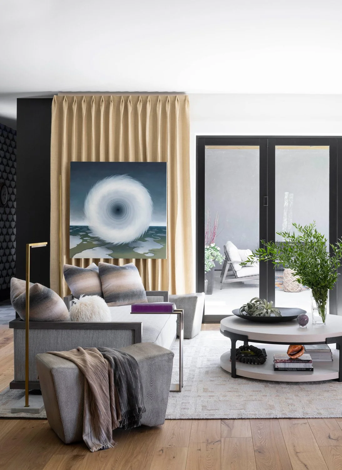 Modern living room with gray sofa, abstract artwork on cream curtain, glass sliding door leading to balcony with outdoor seating, potted plants, and decorative pillows.