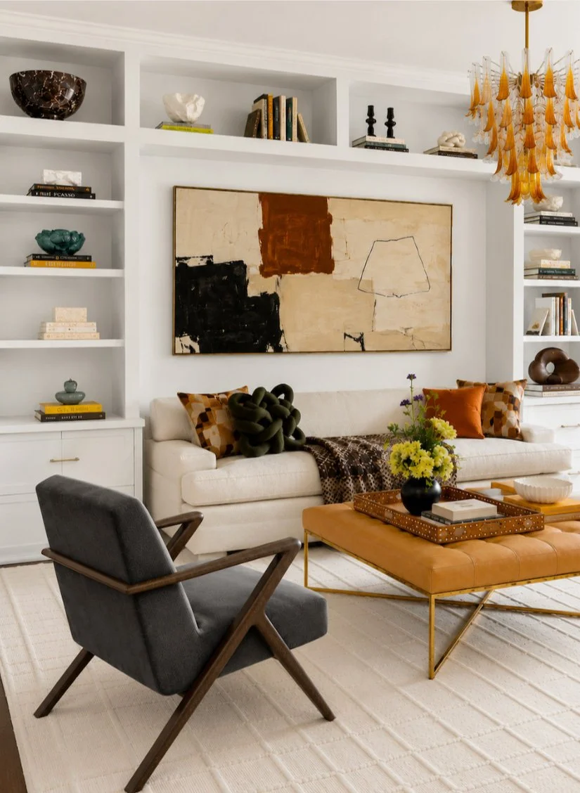 Modern living room with white built-in bookshelves, abstract wall art, beige sofa with patterned pillows, dark gray armchair, orange upholstered coffee table with decorative tray, yellow flowers, and a decorative chandelier.