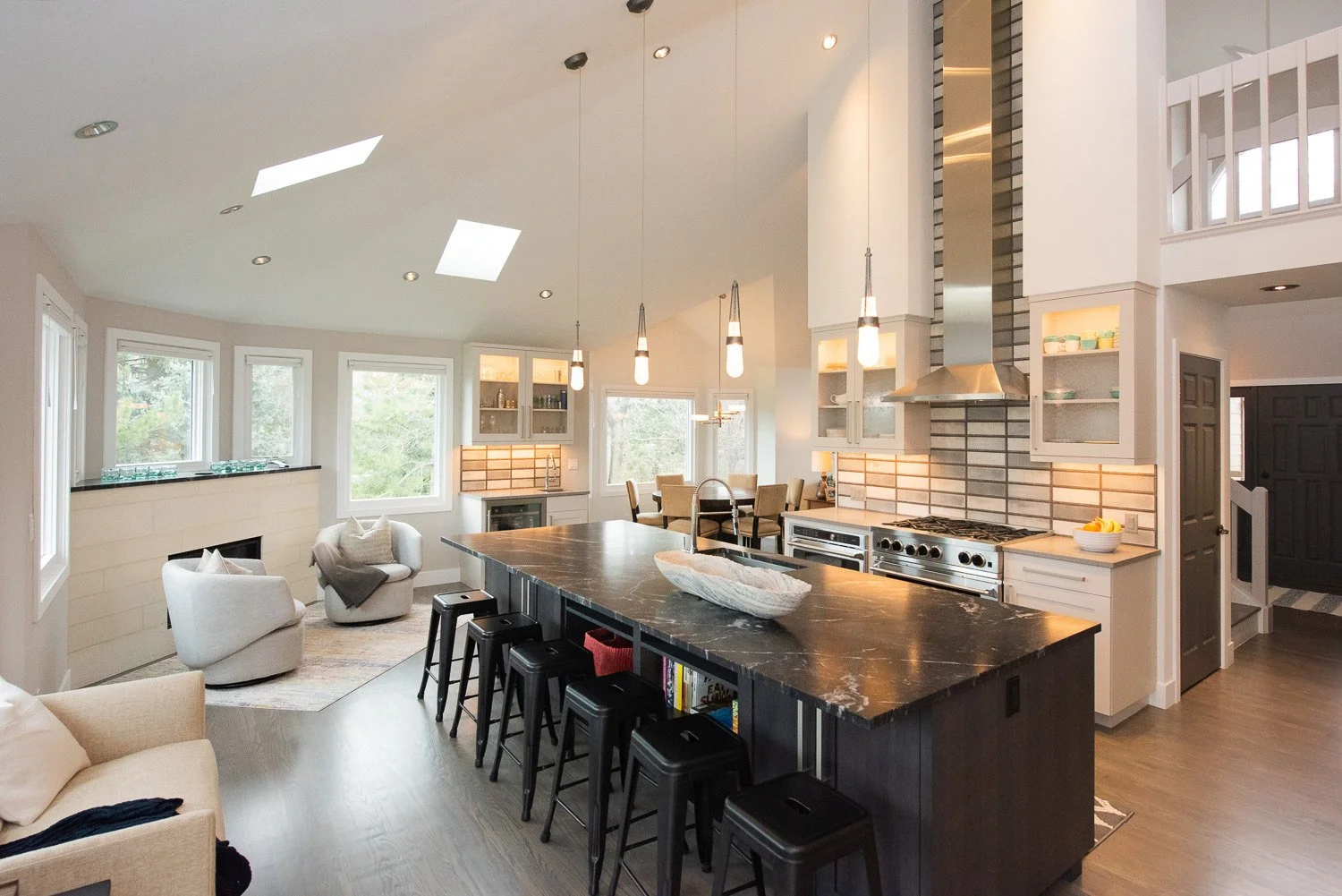 Open-concept kitchen and living room with high vaulted ceiling, skylights, and large windows. Modern kitchen features a black marble island with bar stools, stainless steel appliances, and white cabinets. Living area has white armchairs, a sofa, and 